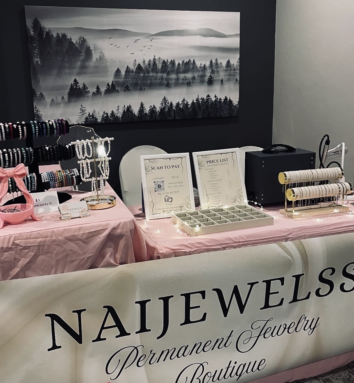 Naijewelss permanent jewelry booth display with pink tablecloth, price list, and chain samples